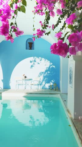 Santorini island, Greece. White cycladic architecture and luxury swimming pool with blue water. Pink flowers over the pool. Travel and vacation concept. Vertical video

