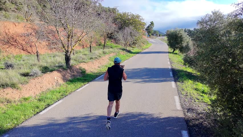 Mature male running in rural surroundings on a sunlit asphalt path, engaging in a cardio workout designed for physical conditioning, mental health benefits, and personal growth