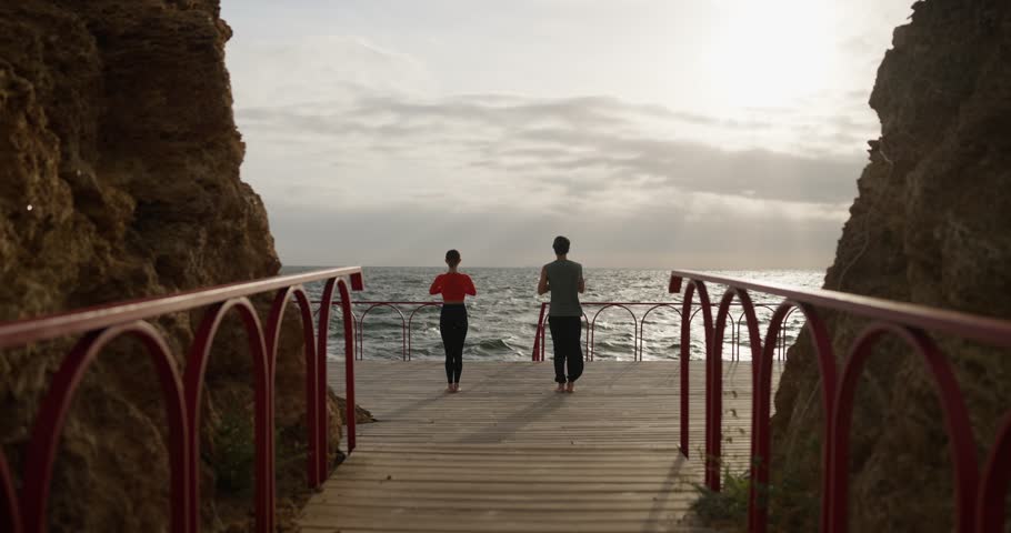 Rear view of a girl together with her boyfriend doing yoga and stretching her leg to the side on the seashore in the summer morning
