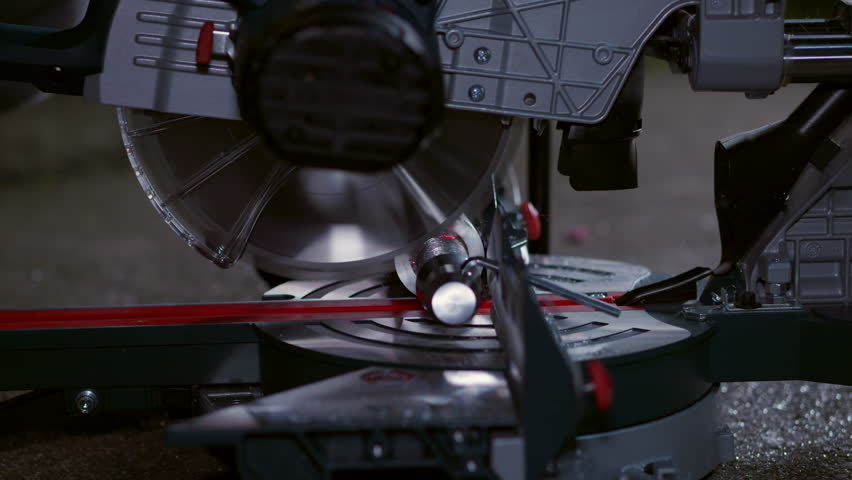 Sawing an aluminum part with a chop saw. Side view of the fast spinning blade used for machining round metal material. Industrial work in a dark outdoor environment. Dust and chips flying around.