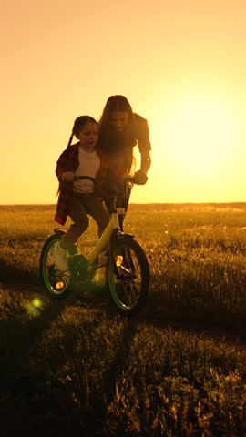 Active Mom teaches her little daughter to ride bike, sunset. Mother teaches her child to keep balance while sitting on bicycle. Childhood dream of riding bike. Family life, mom, baby, parental support