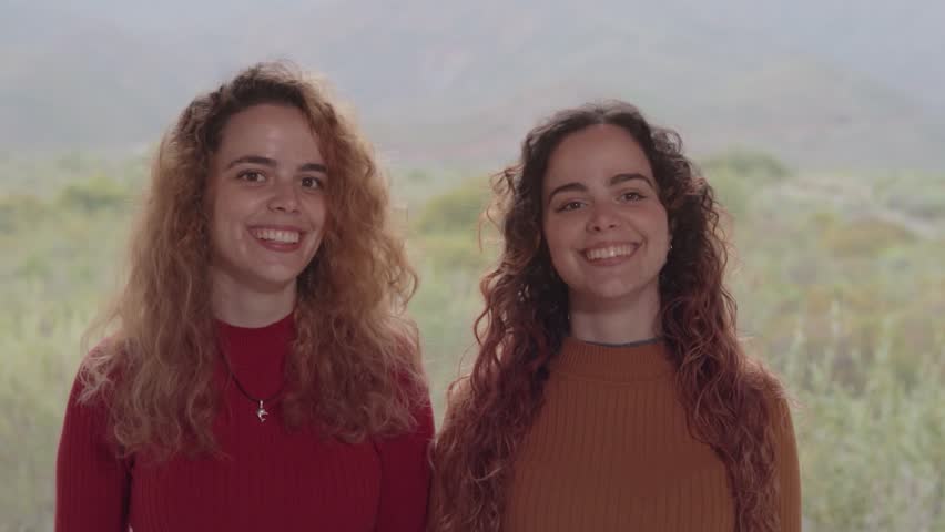 Beautiful Twin Sisters Laughing, Hugging, and High-Fiving in the Green Nature with Mountains in the Background. Hermosas Hermanas Gemelas Riendo, Abrazándose y Chocando la Mano en la Verde Naturaleza 