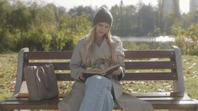 Relaxing on a wooden bench, a woman enjoys a book, surrounded by a calm lake and trees. Her knit hat and beige coat complement the serene autumn setting in the park. - Powered by Shutterstock - Get 15% off with code: PIKWIZARD15