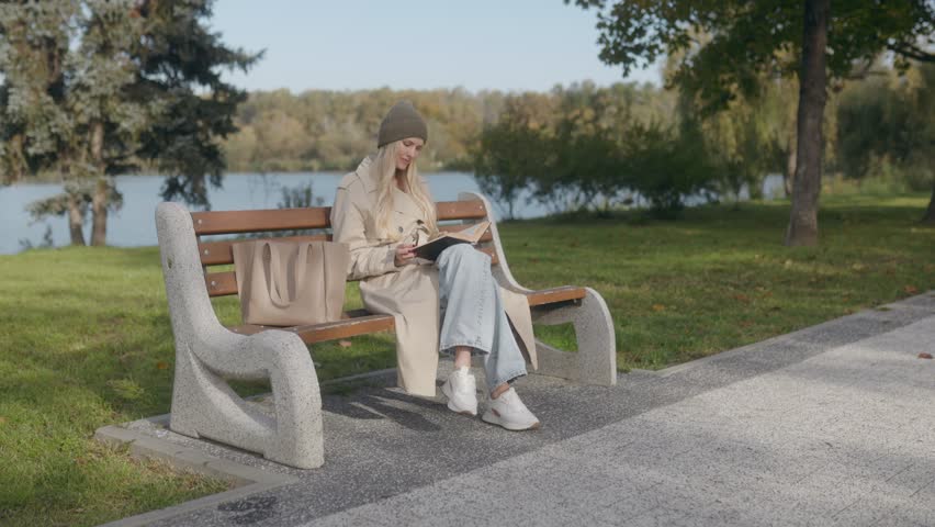 A smiling, blonde woman sits on a bench, reading a book in a scenic autumn park. Dressed for the season, she enjoys the peaceful moment surrounded by natural beauty.