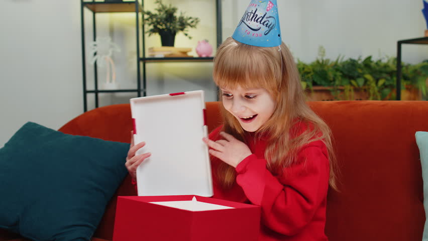 Happy preteen child girl wears festive birthday cap hat hold gift box with ribbon congratulating. Teenager female kid celebrating party event, opening delivery greeting present at home room on sofa