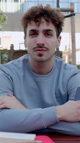 Vertical video. Front view portrait of joyful young student man holding phone smiling at camera sitting on table at campus college. Education and professional creative people concept.