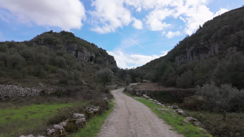 Dirt road winding through a narrow valley with rocky hillsides near Buracas do Casmilo, Condeixa - Portugal. Olive trees, and scattered stone walls under a blue sky with scattered clouds.