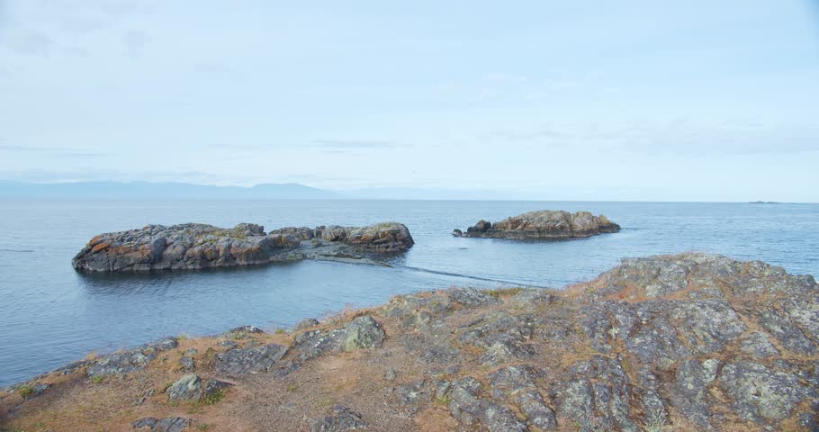 Handheld video of the rocky shoreline at Neck Point Park in Nanaimo, BC, Canada. Calm blue waters stretching to a distant horizon under a cloudy sky. Serene coastal scenery with a sense of tranquility