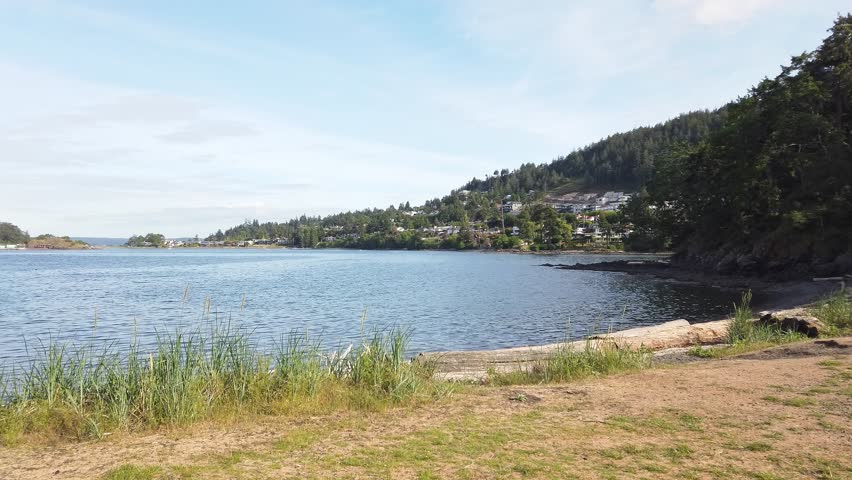 A handheld shot of a tranquil seascape with gentle waves, grassy shoreline, and forested hills near Neck Point Park, Nanaimo, BC, Canada