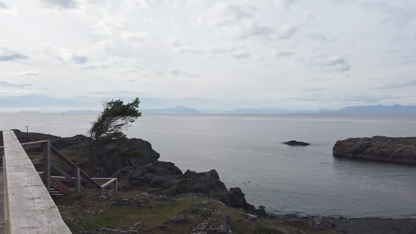 Panning right across Neck Point Park in Nanaimo, BC, Canada, on a clear day. The serene coastal scene features rocky islands, calm ocean waters, and a cloudy sky, with distant mountains on the horizon