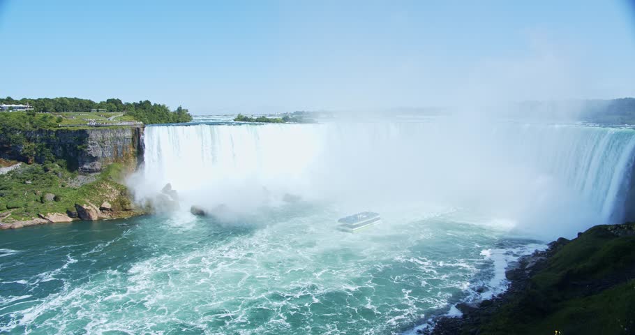 A breathtaking view of Niagara Falls on a sunny summer day, with mist rising as the water cascades down. In the distance, a tour boat approaches the powerful waterfall. A handheld shot.
