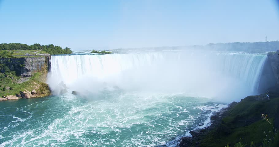 A breathtaking view of Niagara Falls on a sunny summer day, with mist rising as water cascades down. The rushing blue water flows powerfully, creating a serene yet epic scene. A handheld shot