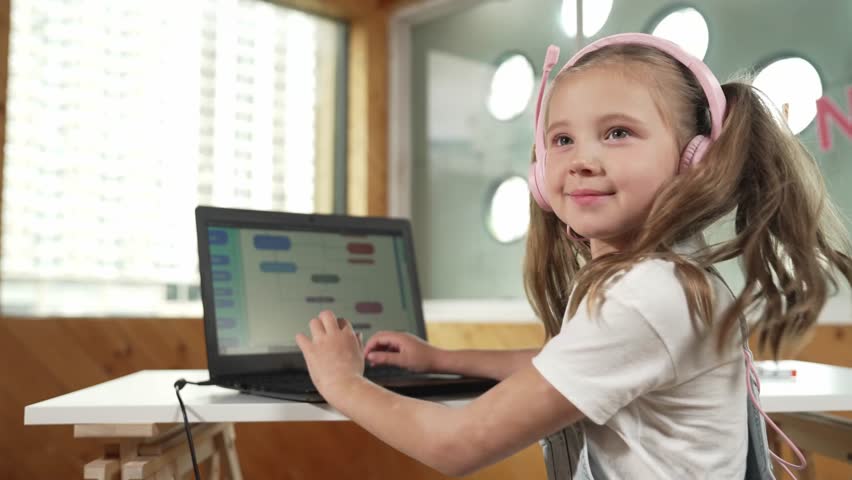Smiling girl using laptop and turn around to show thumb up to camera. Cute child wearing headphone while working by using laptop writing code in STEM technology education. Online learning. Erudition.