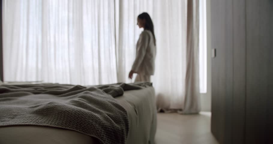 A peaceful bedroom scene with a woman in comfortable clothes standing by the window. The bed features a cozy textured blanket, creating a calm and inviting atmosphere. Relaxing Bedroom
