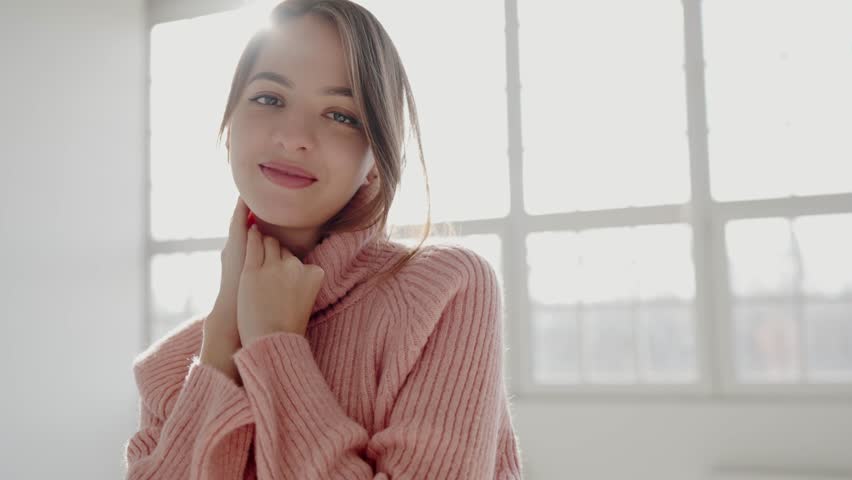 A portrait of a young woman in a warm pink sweater, holding her face softly and smiling. Natural light illuminates the scene, creating a peaceful and cozy ambiance. Elegant Young Woman Posing