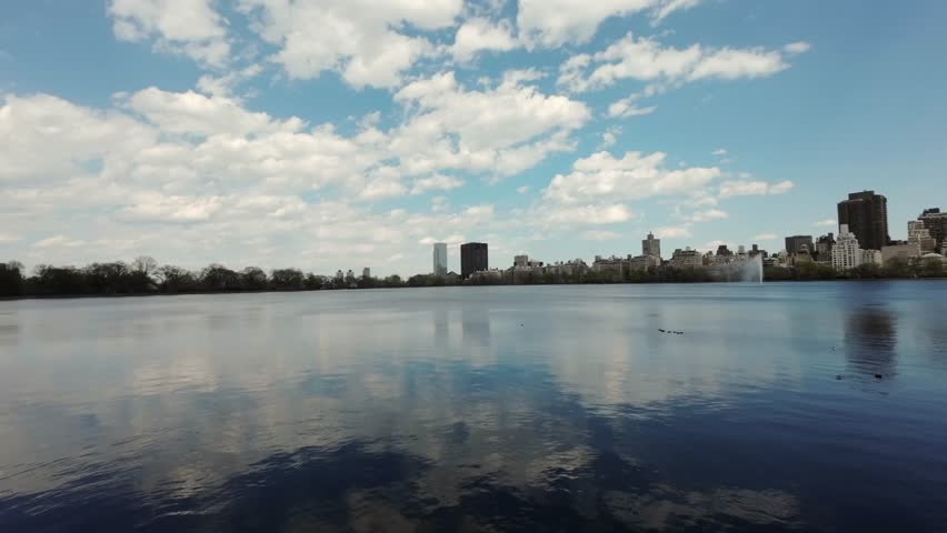 A tranquil urban lake reflects fluffy clouds and a stunning modern city skyline under bright blue skies. New York Central park.
