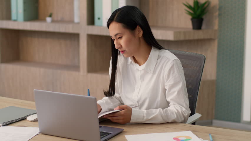 Serious attentive Asian Korean woman female worker Japanese girl company manager businesswoman secretary using laptop video call conference business meeting listening nods head writing notes at office