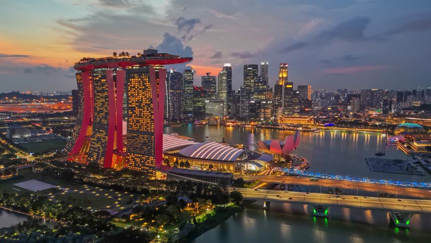 Aerial view of Singapore at dusk. Marina Bay Sands hotel is prominently displayed in foreground. City lit up by streetlights and buildings, reflecting in water of bay. Skyline of Singapore at sunset
