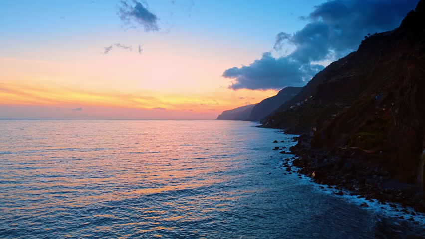 Dark rocks of the Madeira Islands meet the waves of the North Atlantic Ocean. Beautiful pink sky at sunset over the horizon.