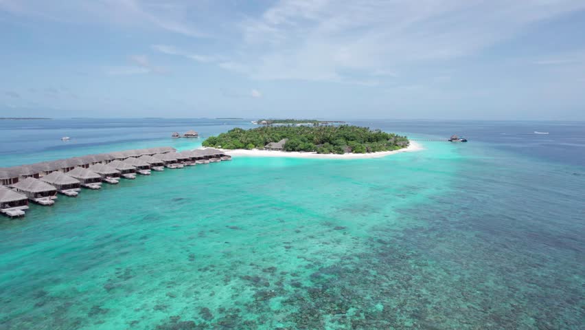 A drone view of an island with white sand in the middle of the ocean. Turquoise water, coral reef. There are bungalows on the water and villas on the beach. There are palm trees all around. Maldives