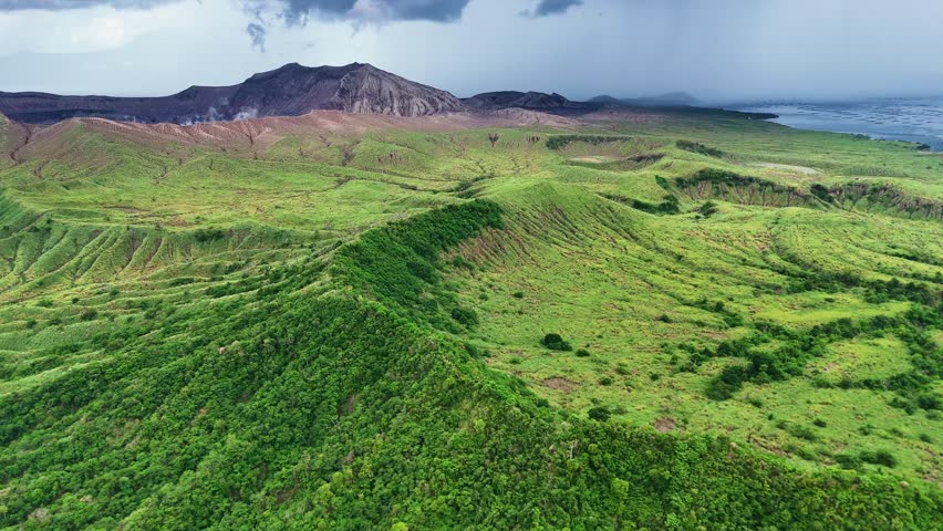 Aerial shot towards Taal Volcano in the Philippines on a cloudy summer day, with thick clouds overhead and distant rain, highlighting the moody, dramatic landscape over Taal Lake.
