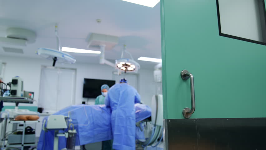 Doors opened to a modern surgery room. Surgeon stands at the operating table his back to camera. Female nurse assisting. Low angle view. Blurred backdrop. - Powered by Shutterstock - Get 15% off with code: PIKWIZARD15