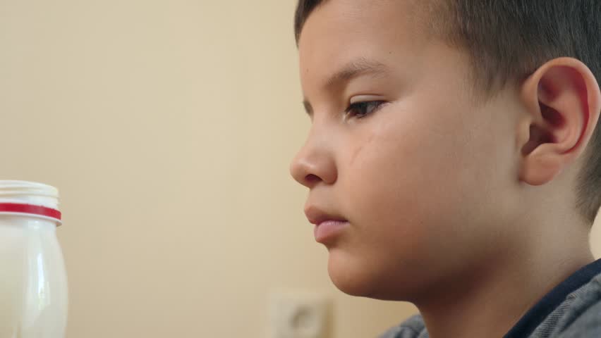The boy drinks yogurt while sitting at the table.