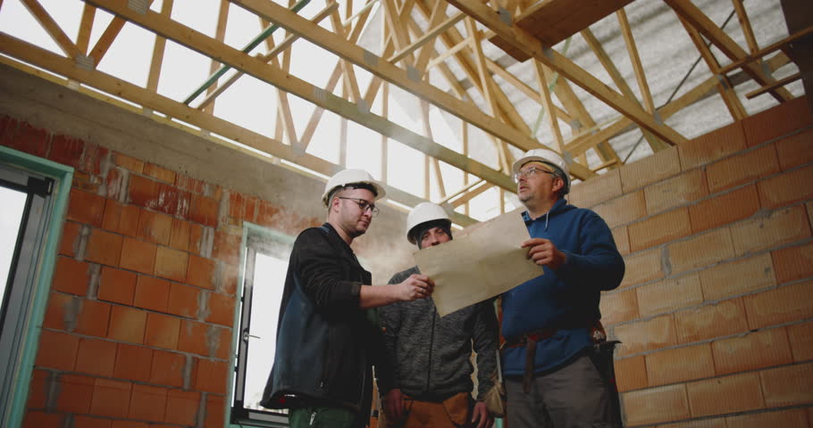 Construction team in hard hats analyzing building plans and pointing at architectural details, collaborating inside an unfinished structure for project coordination