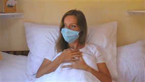 Young woman wearing protective face mask, coughing, resting in bed while holding pillow in bright bedroom. Health and Safety Awareness. - Powered by Shutterstock - Get 15% off with code: PIKWIZARD15
