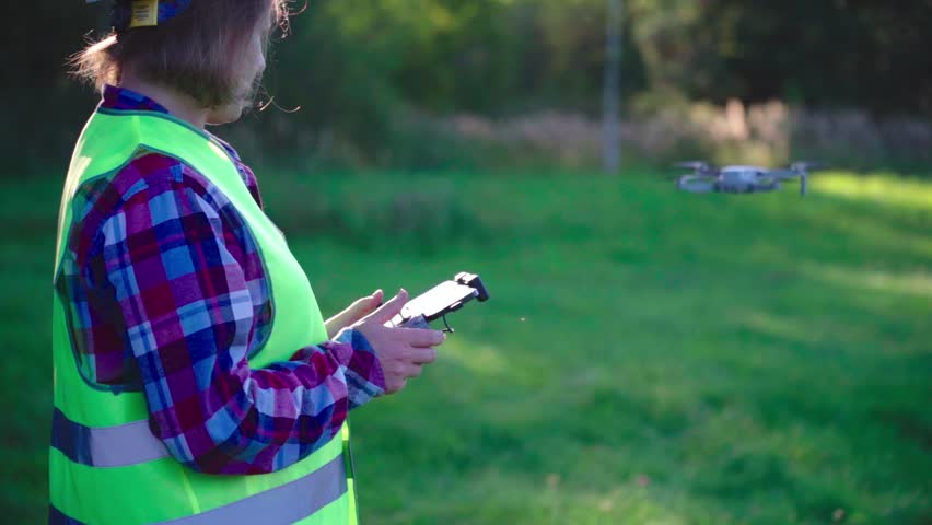 Woman in a yellow vest is holding a cell phone and looking at a drone. The drone is flying in the air above her