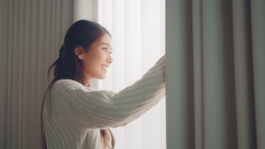Close up of happy young asian woman opening curtain at home in slow motion, Smiling attractive lady looking outside apartment enjoying dreaming and contemplating feeling hope concept