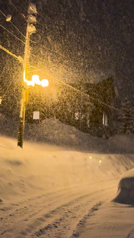 VERTICAL: Dim street lamp casts warm glow over snow laden street during heavy snowfall. Snowflakes dance in the light and create a serene winter scene. Fairy tale with large amounts of snow in Japan.