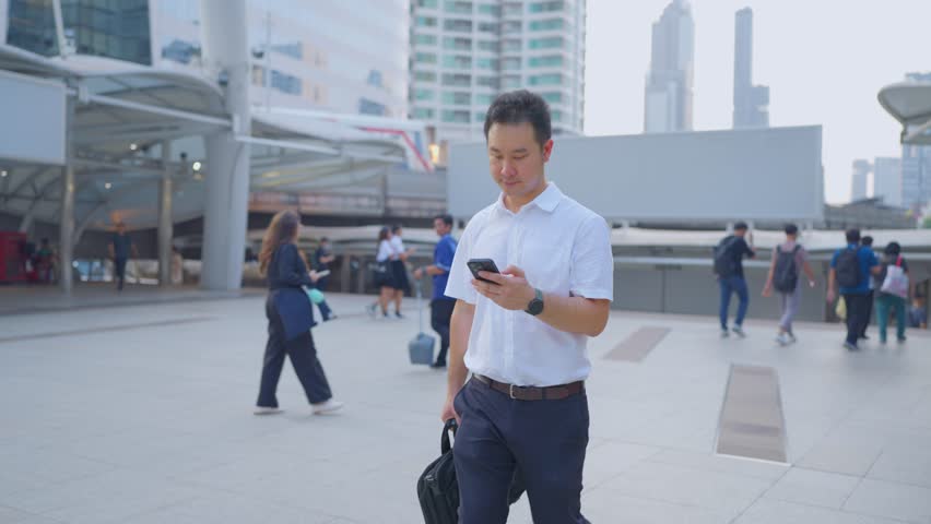 Asian young businessman talking on smartphone while walk in the city. Attractive handsome male employee worker communicate and discuss on mobile cellphone with colleagues outdoors with confidence.

