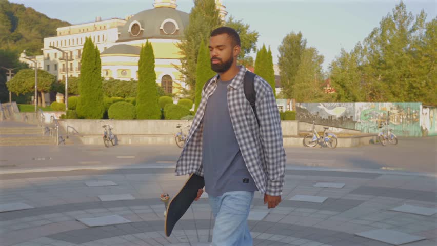 Positive attractive bearded African American man skateboarder with backpack holding skateboard, walking along city street, ready for skateboarding while enjoying recreational pursuit and freedom.