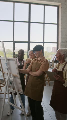 Vertical view of positive female art teacher painting on canvas while three cheerful seniors watching and commenting