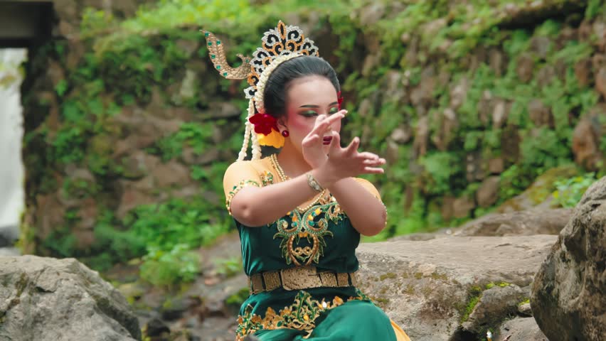 A woman performing a traditional dance in a lush green outdoor setting. She wears a colorful costume adorned with intricate patterns and accessories, showcasing cultural heritage.