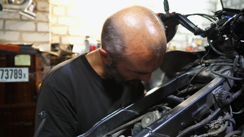 Adult repairman fixing motor of motorbike at workshop. Professional mechanic repairing engine of motorcycle at workplace. Male repairer mending detail of moto at garage. Concept of maintenance service