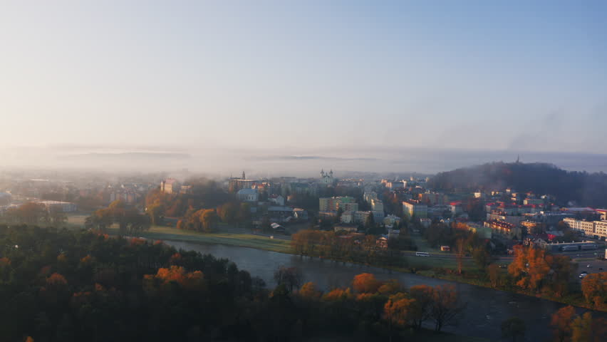 Drone shot of small town covered in early morning fog, buildings and trees bathed in soft, warm sunlight. Mist creates a dreamy effect over the landscape, blue sky above adds a sense of calmness.