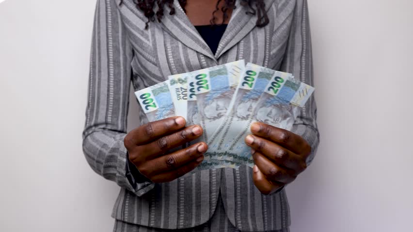 young Black African business woman lady female wearing gray suit standing counting Brazilian Real notes against white background