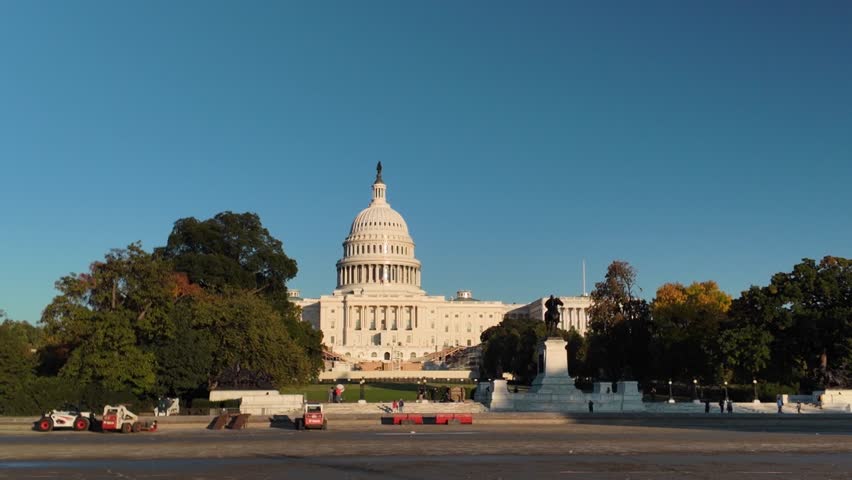 A wide shot of the US Capitol building in Washington DC, with a clear blue sky overhead. Trees line the front of the building, and people walk on the grounds in front. The Capitol building is a beauti