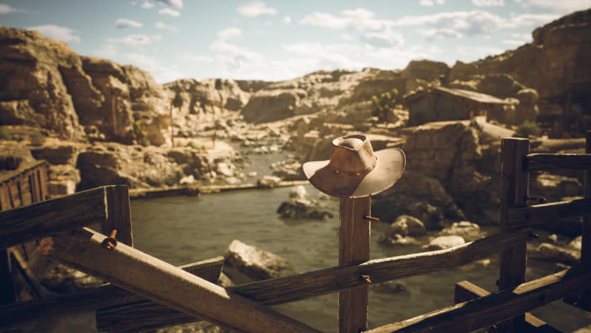 A rustic setting features a cowboy hat placed atop a wooden fence overlooking a serene river. Rocky cliffs and a clear blue sky enhance the tranquil atmosphere of this location.