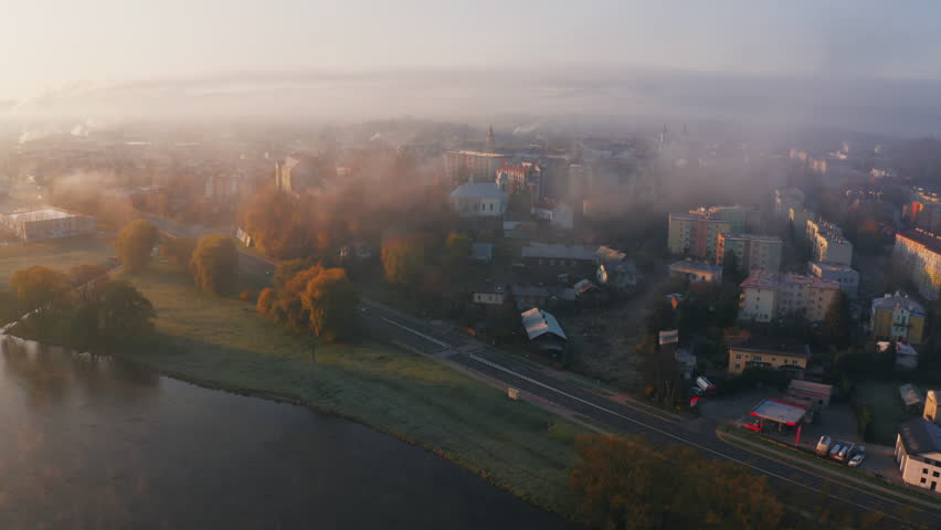 An aerial view of a misty town at sunrise, fog hovering over buildings and a river winding through the landscape in the foreground, creating a vivid, unique, autumn morning scene. Sanok, Poland