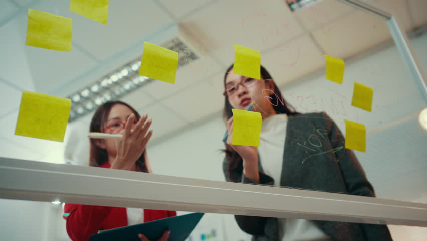 Two businesswomen engage in a project discussion, utilizing sticky notes on a glass wall to brainstorm and organize ideas effectively. Collaboration fosters creativity and strategy