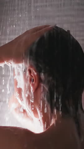 Woman washes hair under shower sprays closeup. Bare lady removes foam from head with water flow in bathroom. Woman enjoys body washing at home