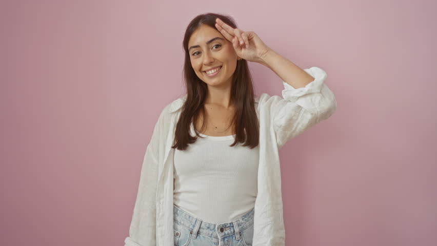 Hispanic woman smiling with peace sign against pink wall, showcasing casual attire and brunette hair in vibrant setting.
