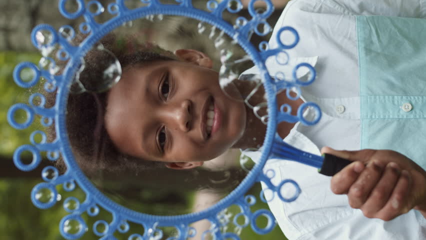 Vertical POV portrait of little African male school kid standing outside, looking and smiling on camera, blowing soap bubbles, holding big wand in hand