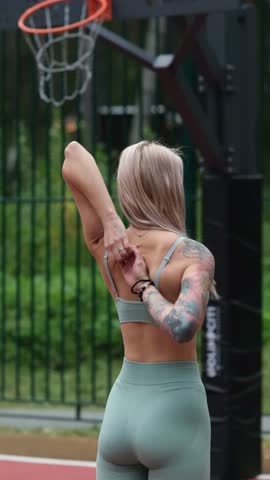 Sporty happy young blonde woman in sportswear doing arm stretching exercises standing on basketball court outside in the morning. Genuine candid photo.