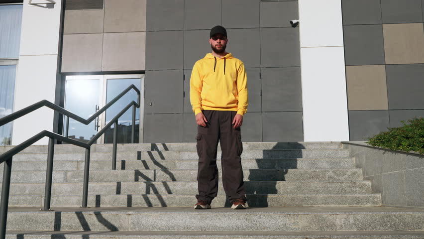 Young dancer man in vibrant yellow hoodie black cap dances confidently on concrete steps in urban environment. Modern building backdrop and casual style reflect street dance vibe in city life. Freedom