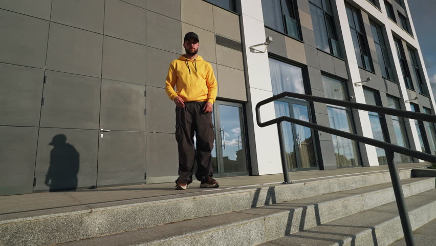 Fanny dancer man in vibrant yellow hoodie black cap dances confidently on concrete steps in urban environment. Modern building backdrop and casual style reflect street dance vibe in city life. Freedom