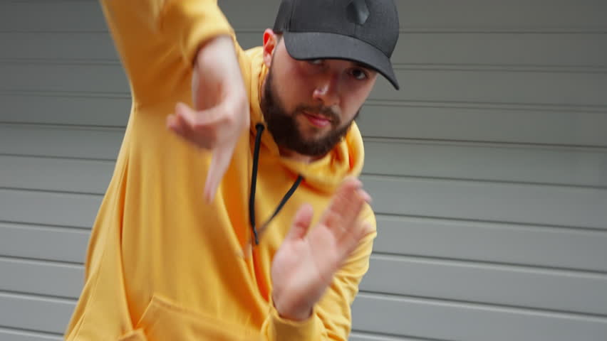 Young dancer male captured mid-dance move in yellow hoodie and black cap stands against gray industrial background. Pose urban attire reflect energy vibe of modern street dance culture. Freedom
